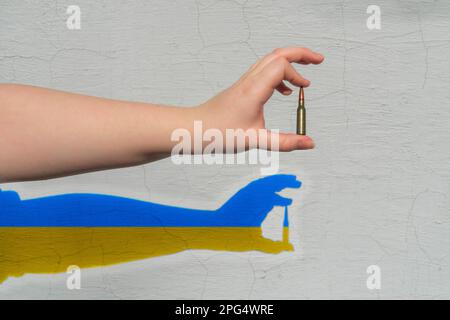 Rifle cartridge in hand. Shadow of yellow-blue color (national symbols of Ukraine) is formed on the wall. The concept of military support for Ukraine Stock Photo