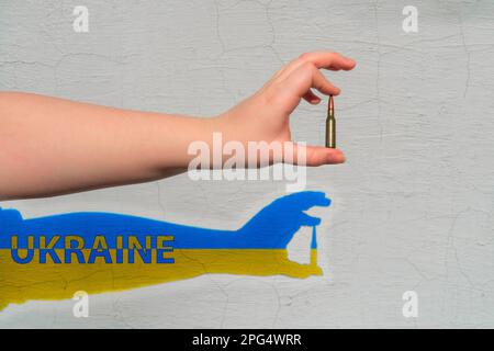 Rifle cartridge in hand. Shadow of yellow-blue color with the inscription - Ukraine is formed on the wall. The concept of military support for Ukraine Stock Photo