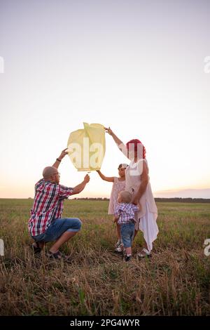 Diversity family launch yellow sky lantern in field at sunset. Mother, father holding Chinese paper lantern, daughter, son wave hands. Making wishes f Stock Photo