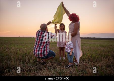 Diversity family launch yellow sky lantern in field at sunset. Mother, father holding Chinese paper lantern, daughter, son wave hands. Making wishes f Stock Photo