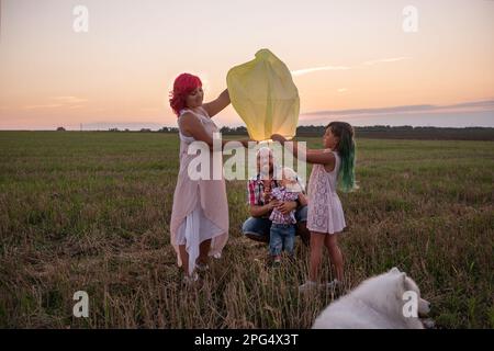 Diversity family with Samoyed dog launch yellow sky lantern in field at sunset. Mother, father holding Chinese paper lantern, daughter, son wave hands Stock Photo