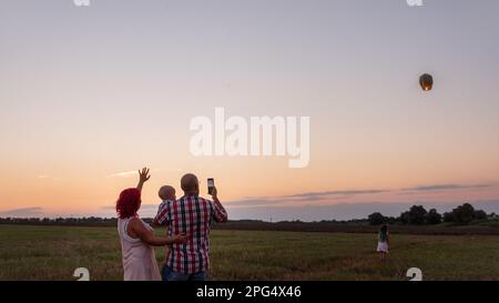 Diversity family taking photo with smartphone yellow sky lantern in field at sunset. Mother father holding little son, daughter wave hands. Making wis Stock Photo