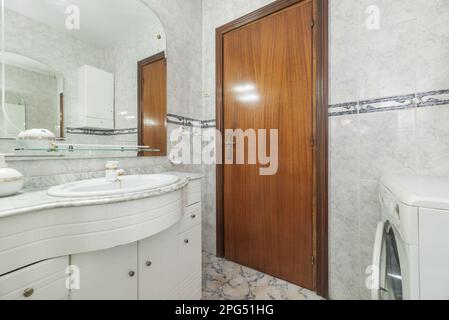 An antique bathroom with a white wooden cabinet with drawers, a porcelain sink with a marble top, a mirror with beveled edges and a washing machine in Stock Photo