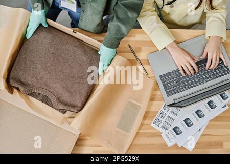 Top view of hands of two volunteers typing on laptop keyboard and packing warm knitted sweater in box by workplace in storage room Stock Photo