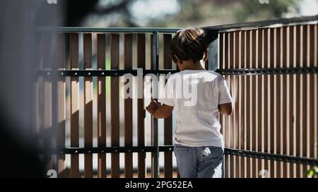 One thoughtful child standing at balcony looking out from second floor. Pensive child holding into security wooden fence at residential building terra Stock Photo