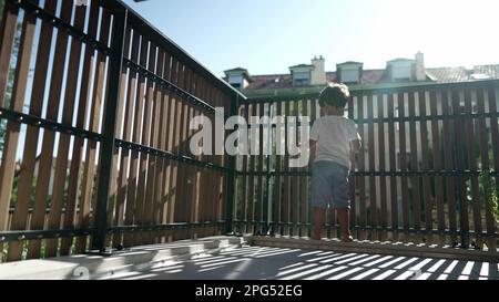 One small boy standing by balcony fence staring outside from second floor with lens flare sunlight. Child holding in wooden bars looking out Stock Photo