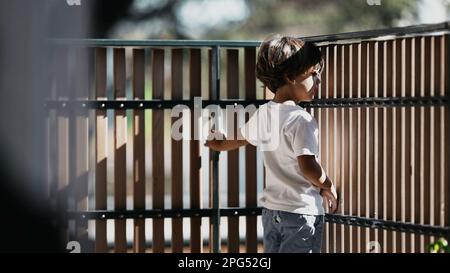 One thoughtful child standing at balcony looking out from second floor. Pensive child holding into security wooden fence at residential building terra Stock Photo