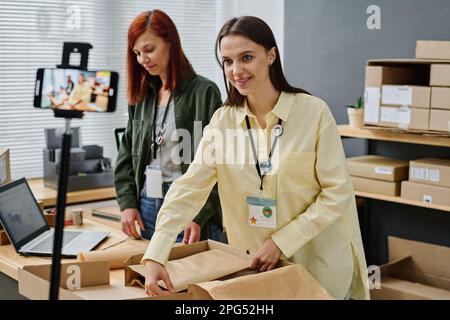 Happy young female volunteer looking at smartphone camera while packing donation box with folded clothes for people in need Stock Photo