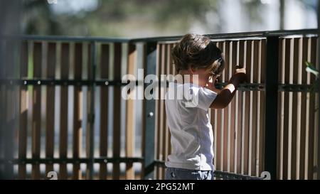 One contemplative small boy standing at balcony holding into wooden fence at second floor of apartment residential home. Pensive child staring out Stock Photo