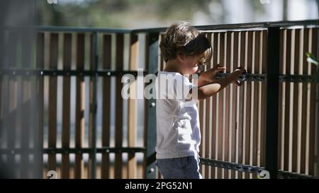 One contemplative small boy standing at balcony holding into wooden fence at second floor of apartment residential home. Pensive child staring out Stock Photo