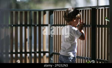 One contemplative small boy standing at balcony holding into wooden fence at second floor of apartment residential home. Pensive child staring out Stock Photo