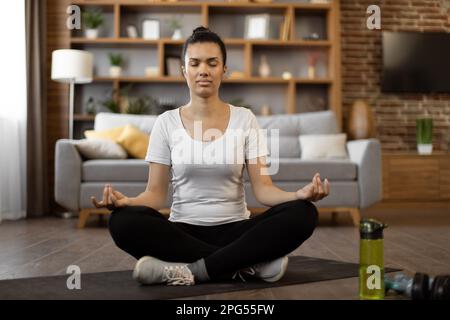 Portrait of african young woman in activewear sitting in lotus position with mudra gesture and smiling at camera. Happy beautiful female meditating at cozy apartment during free time. Stock Photo