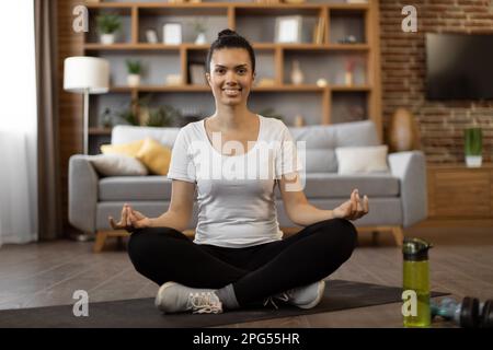 Portrait of african young woman in activewear sitting in lotus position with mudra gesture and smiling at camera. Happy beautiful female meditating at cozy apartment during free time. Stock Photo