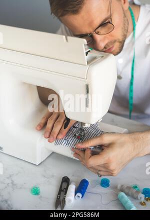 A seamtres with a measuring tape on his neck in a white t-shirt. A blond man in glasses is sewing in home workshop on a sewing machine. The concept Stock Photo