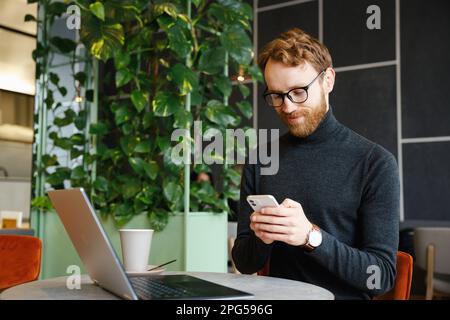 A young red-haired guy, a programmer or businessman in glasses, sits in a stylish cafe at a laptop and chats in a messenger on a smartphone, checks Stock Photo