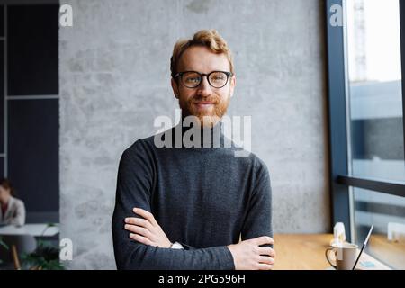 A young red-haired guy, a programmer or an entrepreneur in glasses, in a stylish cafe Looks at the camera and smiles. Freelancer works remotely Stock Photo