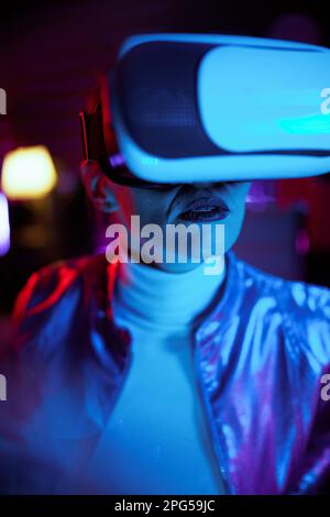 Neon metaverse futuristic concept. stylish middle aged woman in vr headset on dark background. Stock Photo