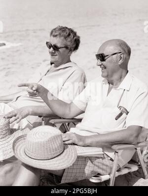 1970s SMILING SENIOR COUPLE MAN AND WOMAN SITTING IN BEACH CHAIRS ON BEACH WEARING SUNGLASSES MAN POINTING - b25261 HAR001 HARS COMMUNICATION SECURITY PLEASED JOY LIFESTYLE SATISFACTION ELDER FEMALES MARRIED SPOUSE HUSBANDS HEALTHINESS COPY SPACE FRIENDSHIP HALF-LENGTH LADIES PERSONS MALES RETIREMENT CONFIDENCE SENIOR MAN GESTURING SENIOR ADULT B&W SENIOR WOMAN SHORE RETIREE HAPPINESS OLDSTERS CHEERFUL OLDSTER LEISURE RECREATION DIRECTION BEACHES GESTURES SMILES ELDERS CONNECTION JOYFUL SANDY STRAW HATS COOPERATION TOGETHERNESS WIVES BLACK AND WHITE CAUCASIAN ETHNICITY HAR001 OLD FASHIONED Stock Photo
