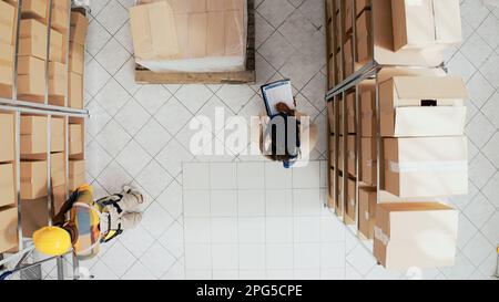 Team of people doing quality control in storehouse, standing between shelves and racks filled with carton packages. Employees working with boxes and merchandise cargo. Drone shot. Stock Photo