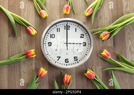 A clock shows three hours. Tulips are lying around. A symbol of the change of time. Daylight saving time. Moving the hands forward. Stock Photo