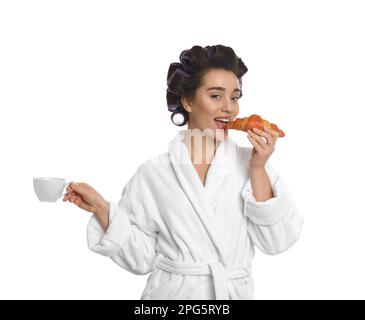 Young woman in bathrobe with hair curlers having breakfast on white background Stock Photo