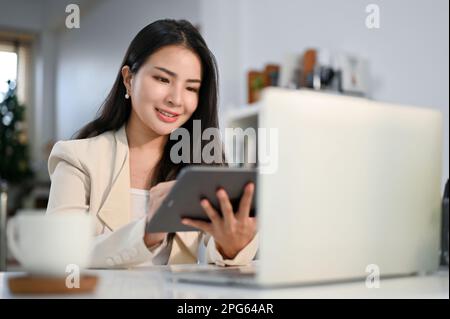 Charming and elegant millennial Asian businesswoman using her tablet, reviewing online documents on her tablet, working at her desk in the office. Stock Photo