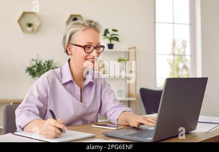 Happy mature woman using her laptop computer for remote work and online education Stock Photo