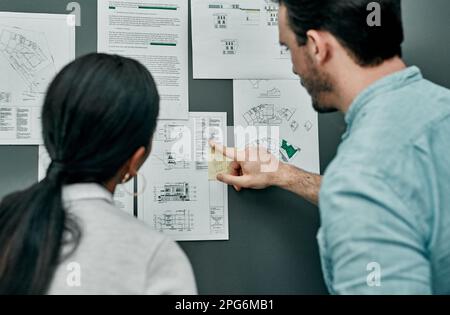 Focusing on the details. Rearview shot of two architects working with blueprints on a wall in an office. Stock Photo