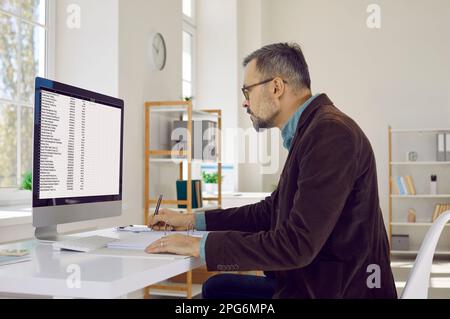 Portrait of serious, attentive, mature businessman man with glasses, sitting at his workplace and looking at his computer screen. Stock Photo