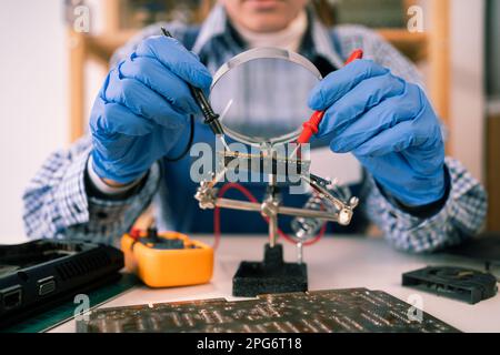 Engineer testing microcircuit through magnifier using multimeter. Electronic renovation in repair shop. Female repairman tinkering computer component Stock Photo