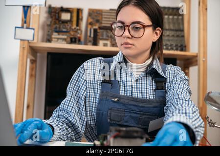 Female computer repairman working on repairing computer in IT workshop watching online courses using laptop. Stock Photo