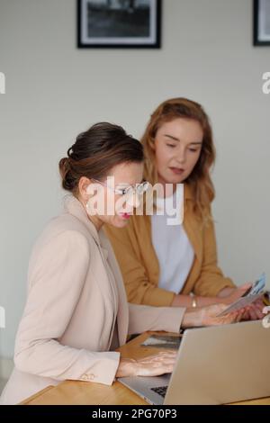 Two female workers of art gallery discussing online information on laptop screen while preparing for new exhibition and choosing pictures Stock Photo