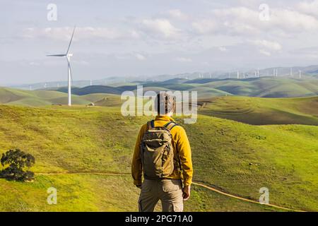 Man looking on the wind power mill among green hills, view from the back Stock Photo