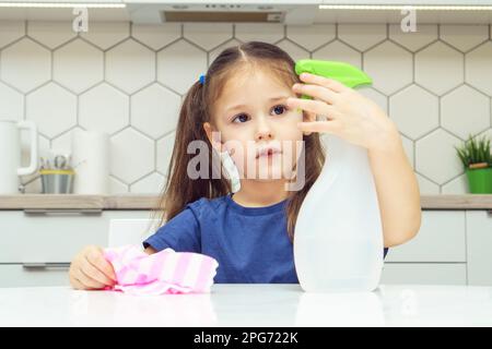 Pretty little girl push lever of detergent sprayer at table. Portrait of child tidying up kitchen with household rag, home cleaning concept. Kid help Stock Photo