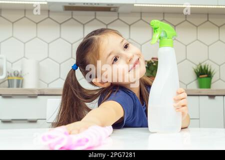 Nice little girl with detergent sprayer and household rag wipe table. Portrait of happy child tidying up kitchen, home cleaning concept. Kid help Stock Photo