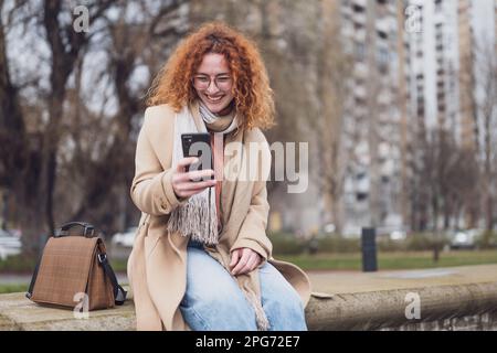 Natural portrait of a caucasian ginger woman with freckles and curly hair. She is smiling and using smartphone. Stock Photo