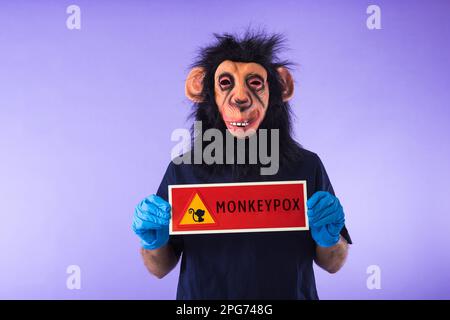 Disguised person with a monkey mask and a doctor's suit, holding a sign with the danger sign and a monkey, which reads: MONKEYPOX, on a purple backgro Stock Photo