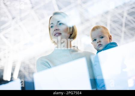 Thoughtful young mother looking trough window holding his infant baby boy child while waiting to board an airplane at airport terminal departure gates. Travel with baby concept Stock Photo