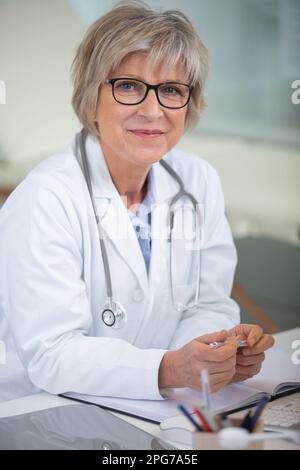 smiling attractive mature female doctor sat at her desk Stock Photo