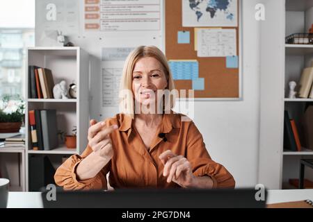 Mature woman sitting at table in front of computer monitor and teaching her students online Stock Photo