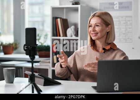 Smiling mature teacher using smartphone for online lesson with her students while sitting at her workplace in classroom Stock Photo