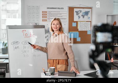 Mature teacher pointing at whiteboard standing in front of camera and having lesson online with students Stock Photo