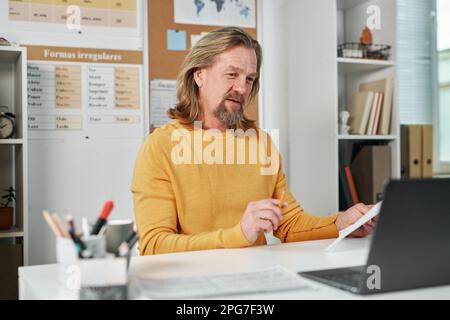 Mature bearded teacher having video call with students on laptop while sitting at his workplace in class Stock Photo