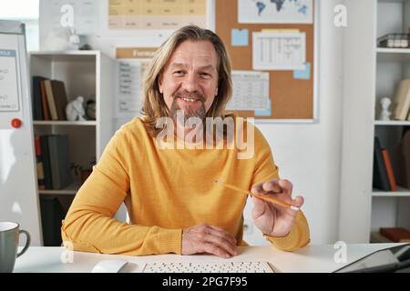 Portrait of mature bearded teacher sitting at table in the class and smiling at camera, he having online lesson with students Stock Photo