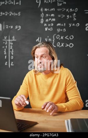 Vertical image of mature teacher sitting at table with laptop and explaining topic for students during online lesson Stock Photo