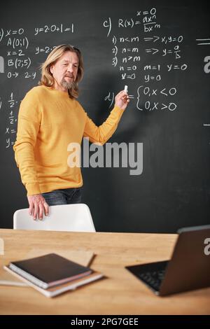 Mature teacher writing new material on blackboard and explaining it to students during online lesson in the classroom Stock Photo