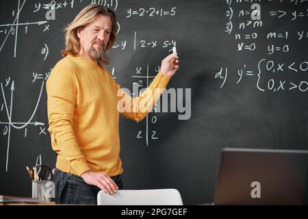Mature teacher standing near the blackboard and explaining Maths to students online using laptop Stock Photo