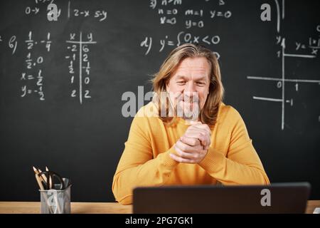 Mature teacher sitting at table in class and using laptop for video call with his students Stock Photo
