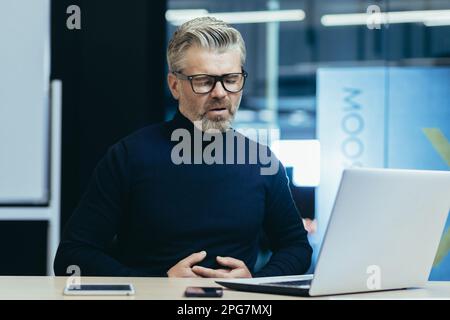 Sick gray-haired businessman poisoned, boss holding stomach, having severe stomach pain mature man working in modern office using laptop. Stock Photo