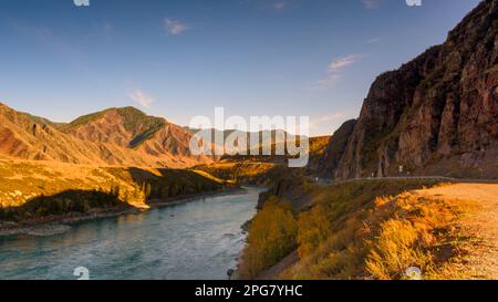The mountain road Chuisky tract goes under a rock in the shade next to the Katun River in Altai in autumn in Siberia during the day. Stock Photo